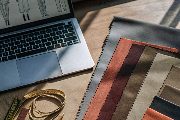 Fabric swatches, measuring tape, and a laptop with clothing sketches on a wooden desk.