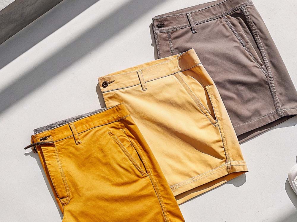 Three pairs of chino shorts in mustard yellow, light beige, and taupe, laid out under bright natural light.