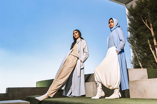 Two women in stylish, modest clothing with soft neutral tones and long coats, posing outdoors against a clear blue sky.