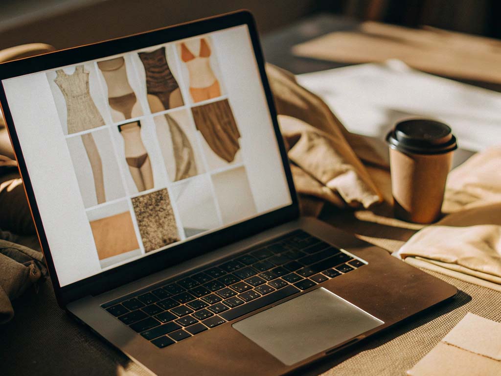 Open laptop displaying swimwear designs and fabrics, with a coffee cup and neutral fabrics nearby.