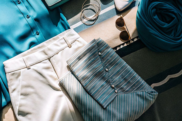 Flat lay of a teal blouse, cream pants, matching clutch, white heels, and accessories, including sunglasses and jewelry, arranged on a striped fabric background.