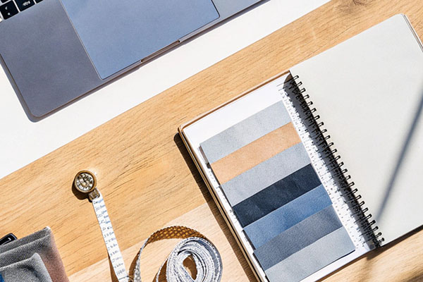 Wooden workspace with a laptop, notebook displaying neutral-toned fabric swatches, measuring tape, and sewing materials arranged neatly under bright light.