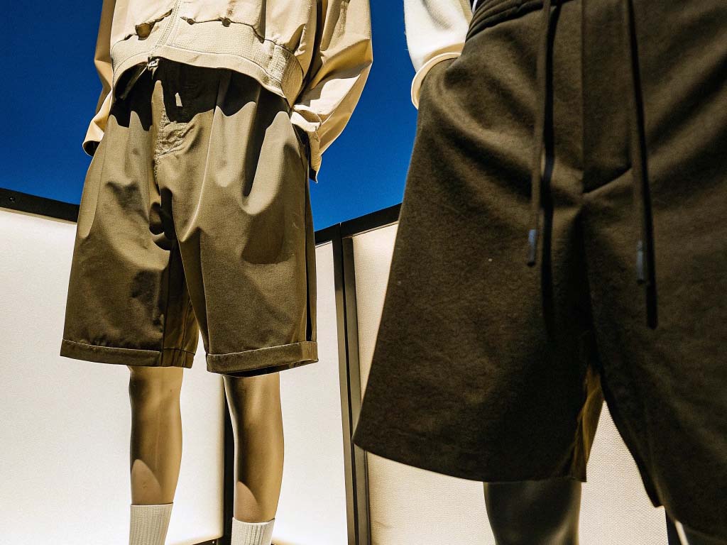 Two mannequins wearing high-waisted belted shorts in tan and red, displayed under warm spotlight in a retail setting.