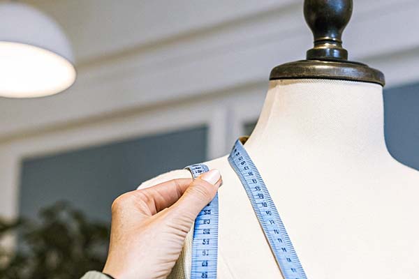 Hand holding a blue measuring tape around the shoulder of a white mannequin in a well-lit studio setting.
