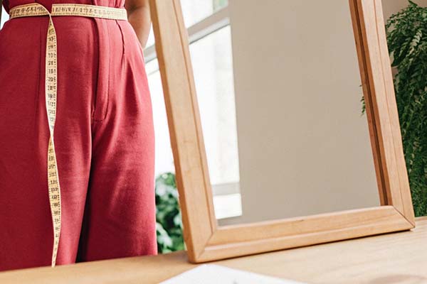 Person in red pants measuring waist using a tape, reflected in a wooden frame mirror.