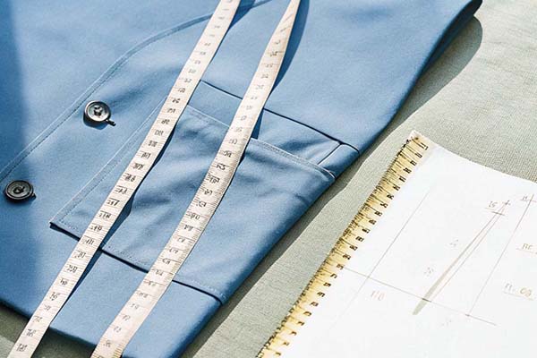 Blue fabric with buttons, measuring tape, and a spiral-bound notebook with pattern sketches, laid on a green surface.