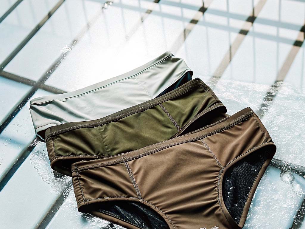 Three pairs of swimwear bottoms in white, olive green, and brown placed on a reflective, water-splashed surface.