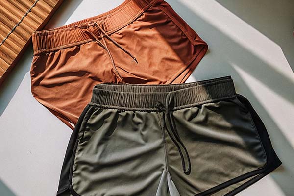Two pairs of athletic shorts in orange and olive green with black accents, featuring elastic waistbands and drawstrings, laid on a white surface under sunlight.