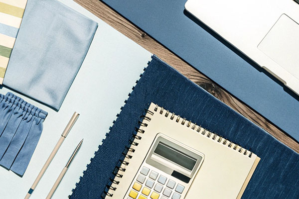 Flat lay of a notebook, calculator, blue fabric swatches, pens, and a laptop on a blue and wooden desk setup.