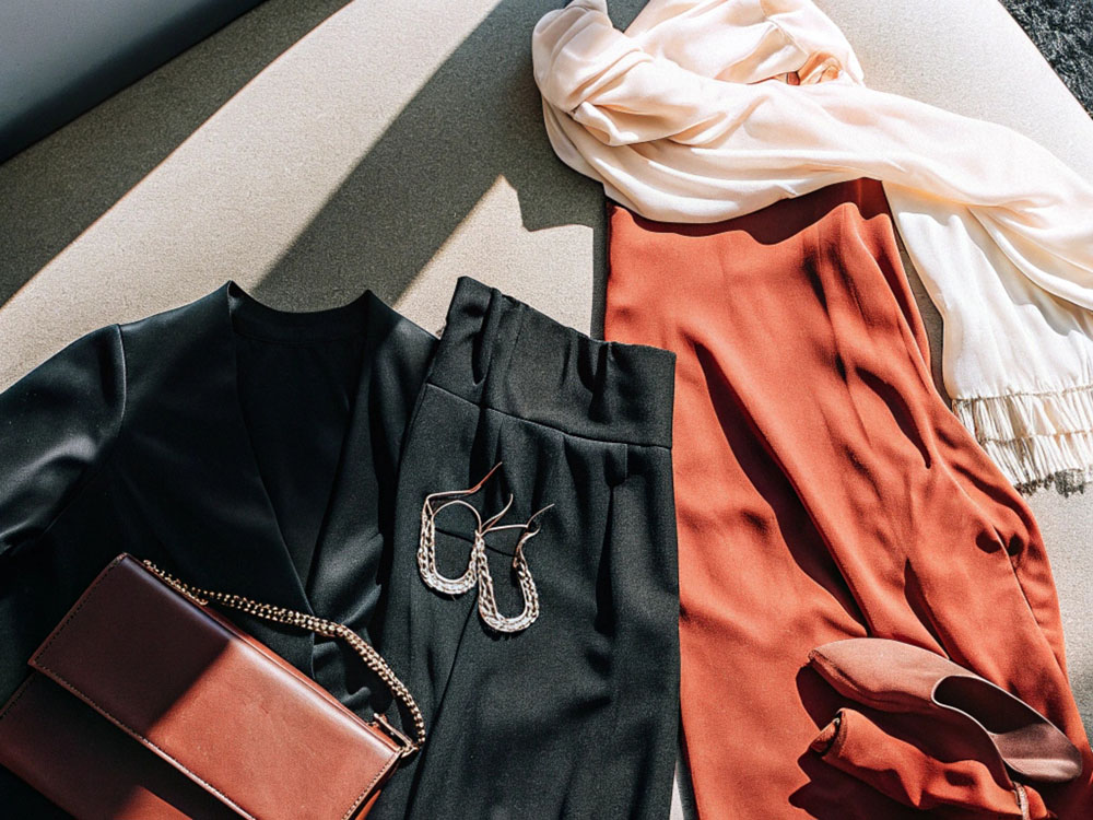 Flat lay of a black top with trousers, a rust-orange dress, a matching bag, scarf, and accessories, including earrings and heels, arranged under natural light.