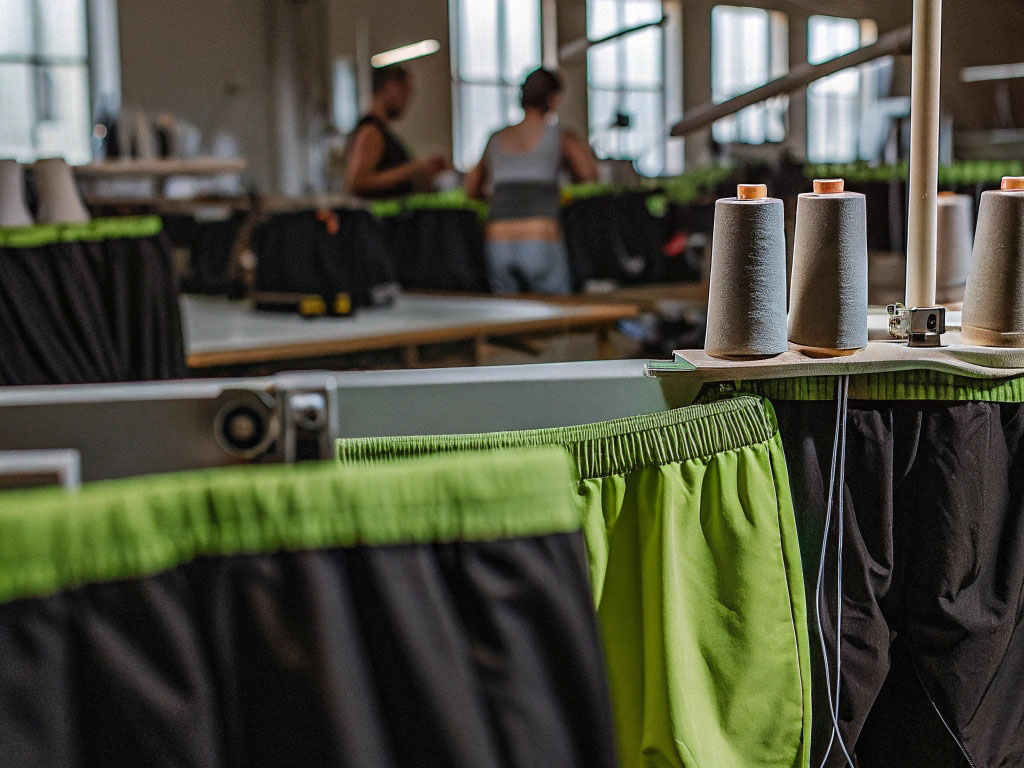 Green and black shorts in a factory setting with sewing machines and fabric spools.