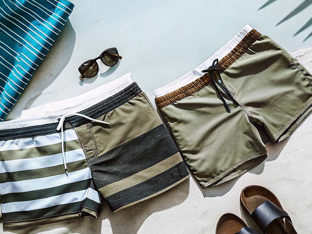 Two pairs of swim trunks, one striped and one solid with a drawstring, styled with sunglasses, sandals, and a striped towel.