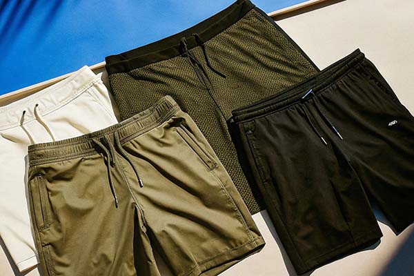 Four pairs of casual shorts in white, olive, textured green, and black, displayed under sunlight.