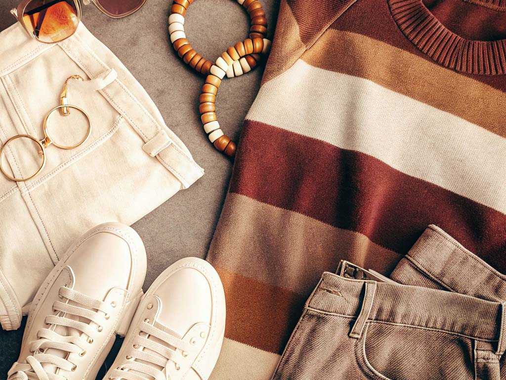 Beige shorts, striped sweater, white sneakers, sunglasses, and accessories arranged on a gray surface.