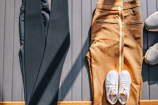 Flat lay of black leggings, tan joggers with zipper details, and white sneakers arranged on a gray surface under natural sunlight.
