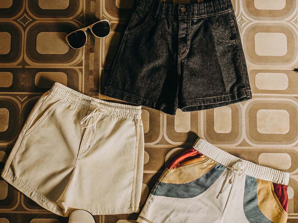 Three pairs of shorts in black, white, and multicolored designs with sunglasses on a retro-patterned surface.