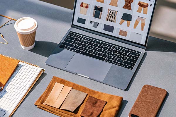 Laptop showing swimwear and fabric designs, with fabric samples, a notebook, coffee, and a phone on a desk.