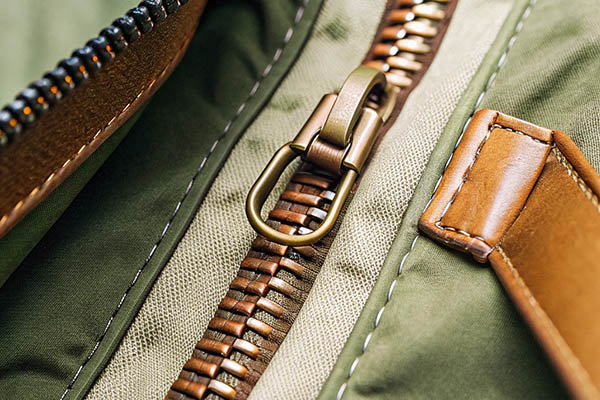 Close-up of a brass zipper on green fabric with brown leather detail and stitching.