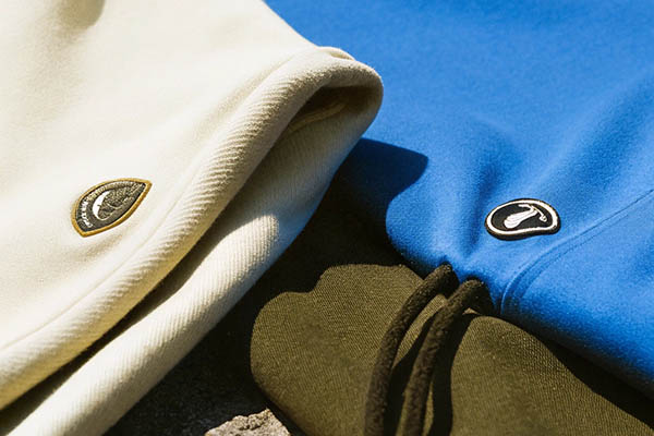 Close-up of cream, blue, and green hoodies with embroidered logo patches in sunlight.