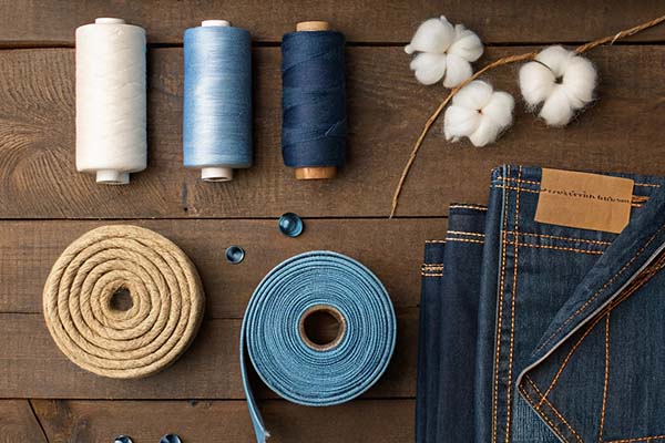 Spools of blue and white thread, cotton bolls, denim fabric, and sewing materials arranged on a wooden surface.