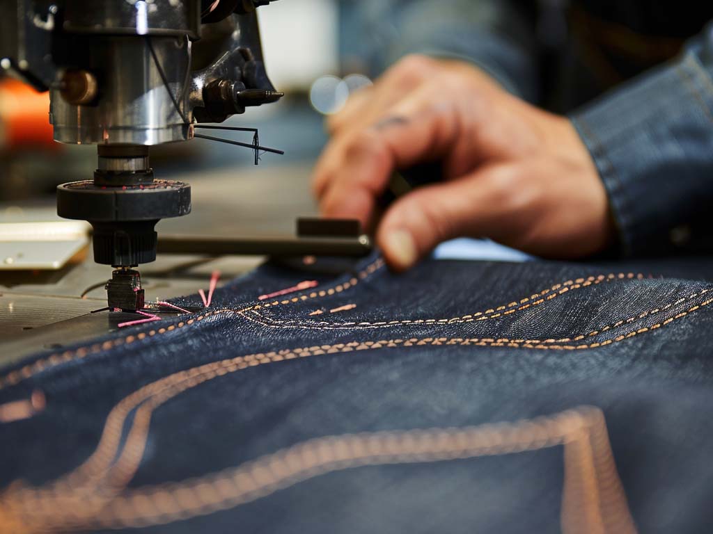Close-up of a sewing machine stitching detailed patterns on dark blue denim fabric with orange thread, guided by a worker's hands.