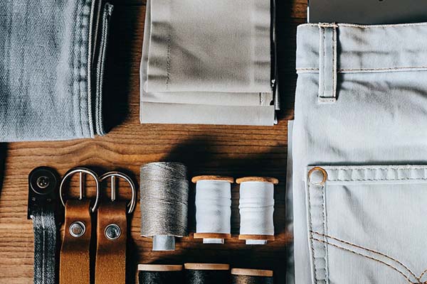 Light-colored denim jeans, fabric swatches, leather straps, spools of thread, and sewing accessories arranged on a wooden surface.