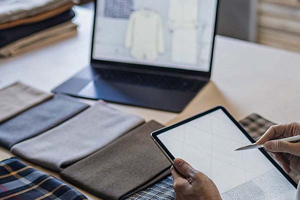 Designer using a tablet to sketch patterns, with fabric swatches and a laptop displaying clothing designs.