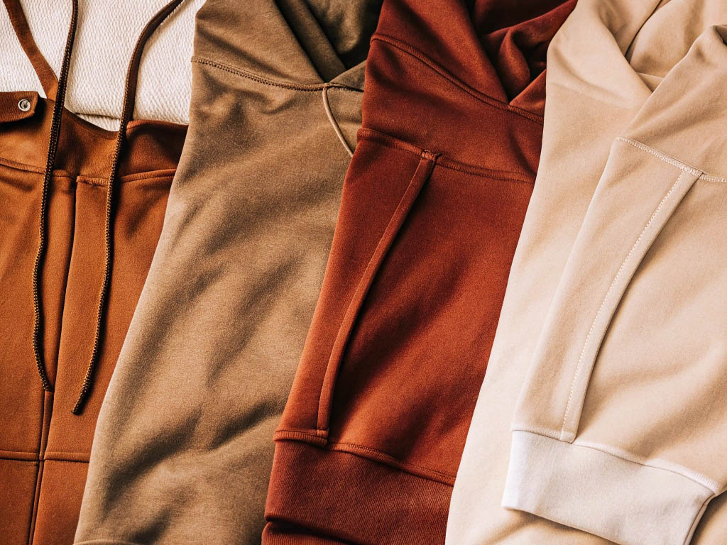 Four hoodies in warm earth tones—brown, olive, rust, and beige—laid flat with soft textures.