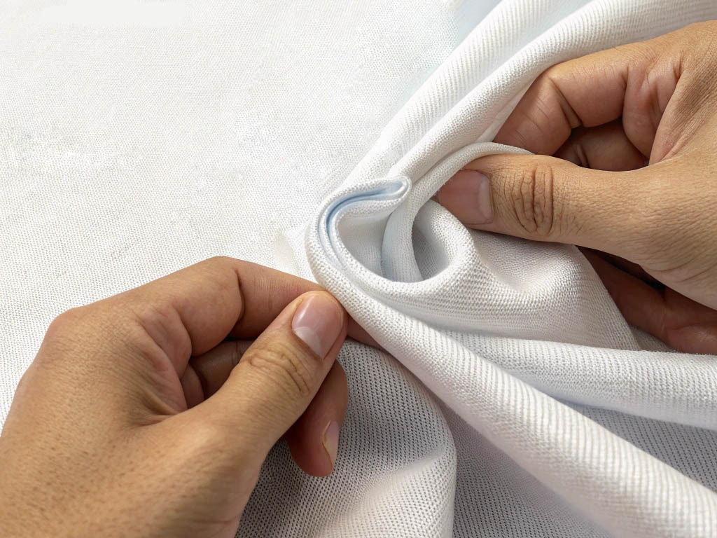 Hands stretching a white fabric to test its elasticity, durability, and softness, with quality assurance icons.