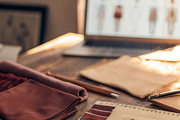 Cozy fashion design desk with fabric samples, sketches, and a laptop displaying clothing designs.