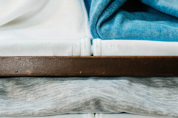 Neatly folded cotton fabrics in white, blue, and gray, stacked on a wooden shelf.