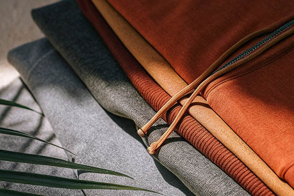 Stacked hoodies in gray, mustard, and rust tones with drawstrings, highlighted by natural sunlight.