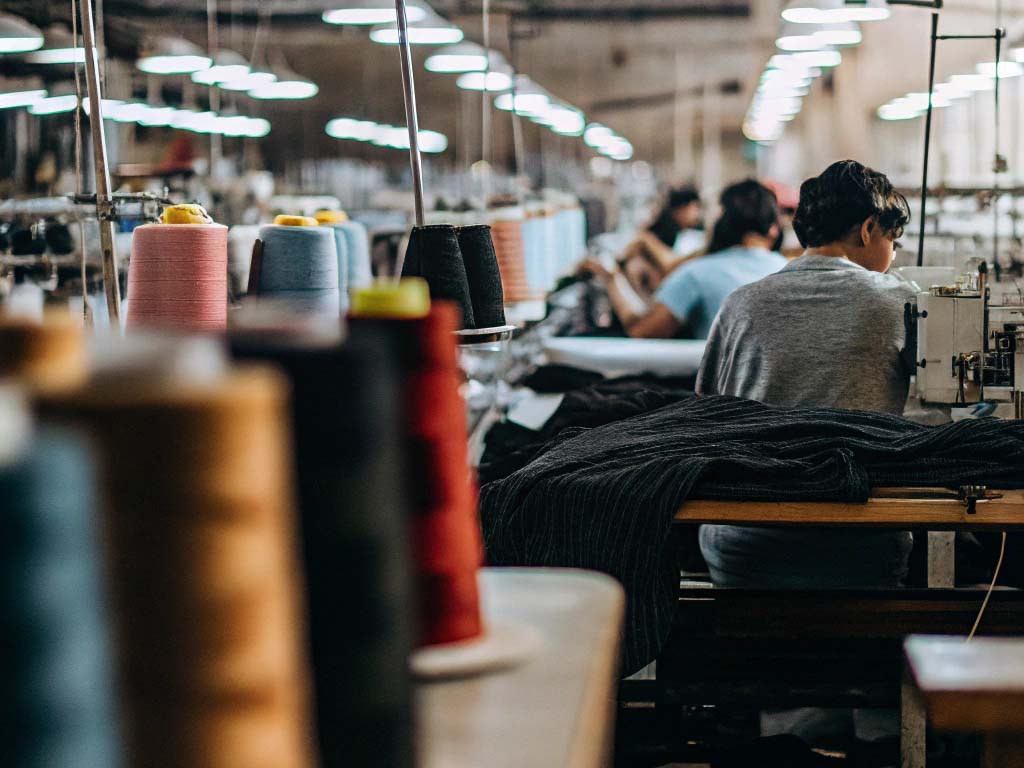 Workers sewing fabrics in a busy garment factory with spools of thread and sewing machines.
