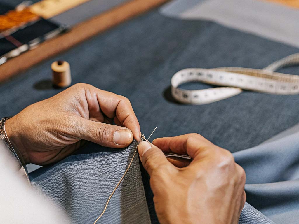 Close-up of a tailor's hands stitching fabric with a needle and thread, measuring tape nearby.