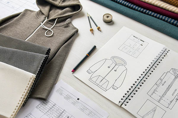 A workspace with hoodie design sketches, fabric swatches, pencils, and a gray hoodie on a white table.