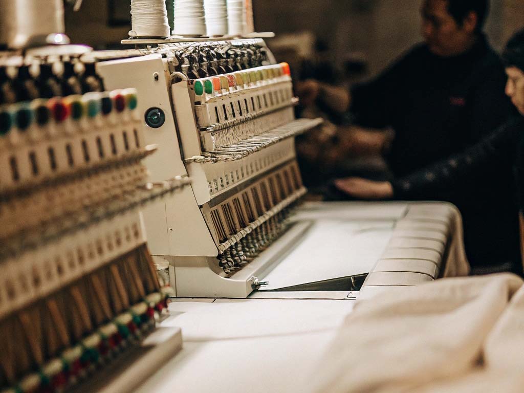 Close-up of an industrial embroidery machine in a textile factory, with workers operating in the background.