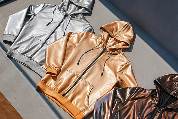Three metallic hoodies in silver, gold, and bronze with a shiny finish, laid flat in sunlight.