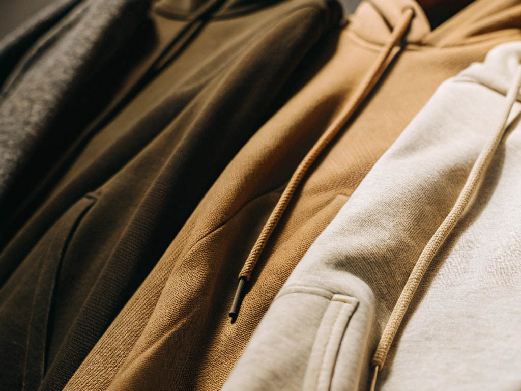 A row of hoodies in neutral shades like gray, olive, camel, and beige, hanging with soft lighting.