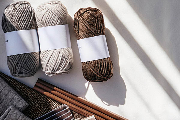 Three skeins of neutral-colored yarn and crafting materials like knitting needles and fabric pieces arranged on a flat surface.