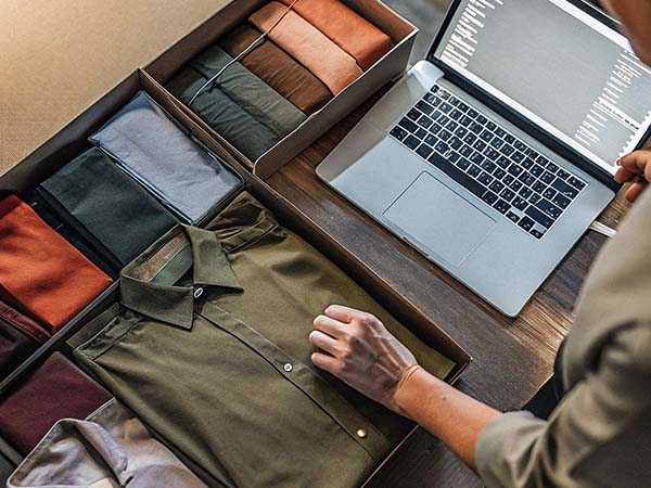 Neatly folded shirts in earthy tones placed in boxes beside a laptop displaying a clothing inventory system.