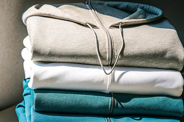A stack of three hoodies in beige, white, and teal, neatly folded in natural sunlight.