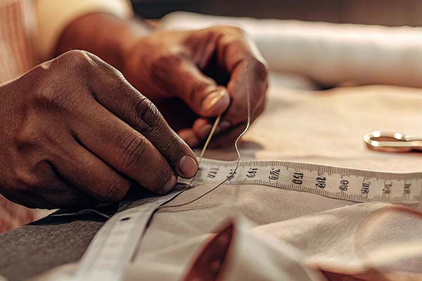 Close-up of a tailor sewing fabric by hand with a needle, thread, and measuring tape.