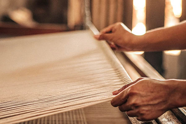Hands carefully weaving fabric on a loom, showcasing traditional textile craftsmanship in warm lighting.