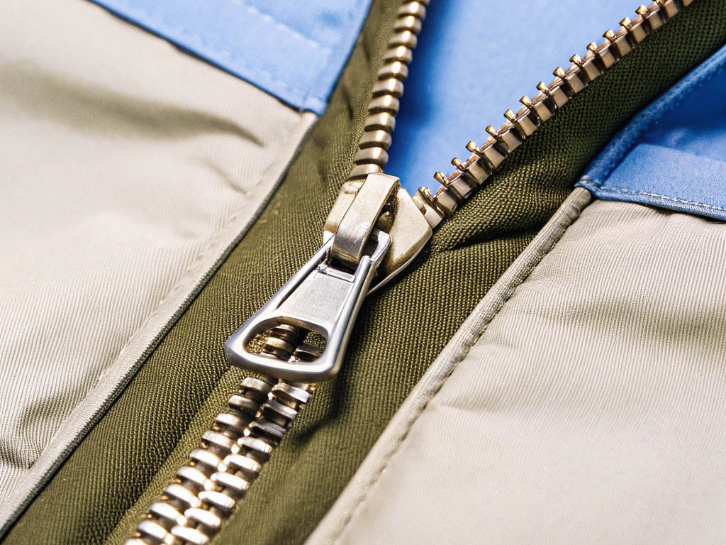 Close-up of a silver metal zipper on beige, green, and blue fabric with stitching.