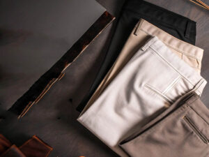 A neatly arranged set of folded chino pants in black, beige, cream, and brown on a dark surface.