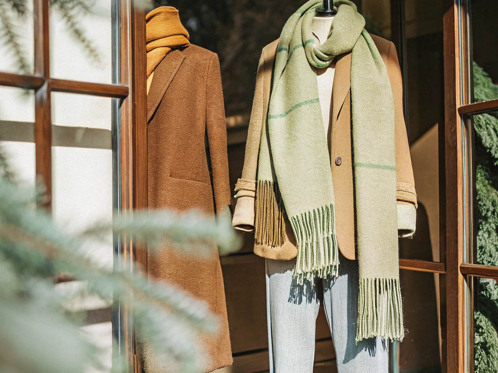 Two mannequins in a store window dressed in stylish camel coats, one with a mustard scarf and the other with a green fringed scarf.