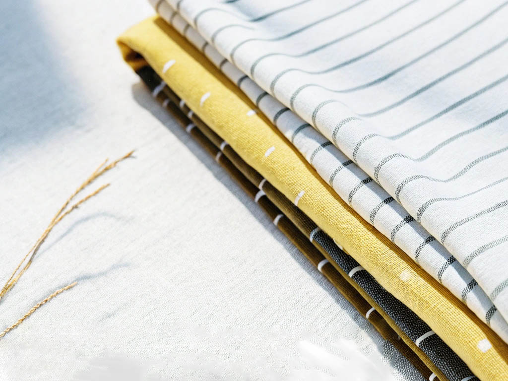Neatly stacked linen fabrics in striped and solid designs, featuring white, yellow, and black tones, bathed in natural sunlight.