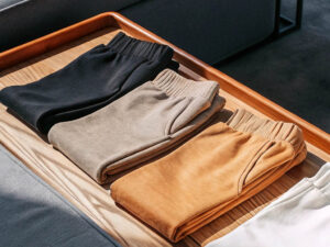 Three neatly folded sweatpants in black, taupe, and camel colors on a wooden tray.