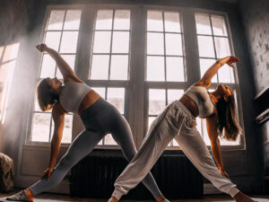 Two women in athletic wear doing yoga stretches in front of large windows.