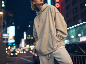 Fashionable woman in an oversized beige hoodie and sweatpants posing confidently in a vibrant cityscape with neon lights and blurred traffic at night.